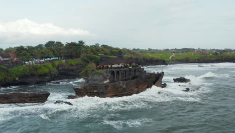 Strong-ocean-waves-crashing-into-rocky-cliff-with-Tanah-Lot-temple-in-Bali,-Indonesia.-Aerial-shot-circling-famous-tourist-destination-at-sea