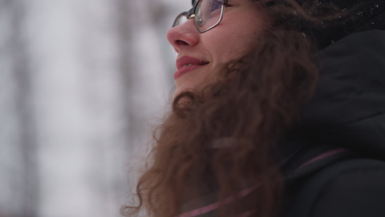 Female wearing glasses and black knitted hat outdoors in snowy environment close up side profile face with curly hair looking upward thoughtful expression winter season blurred forest background