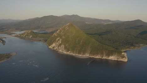 Una-Vista-Del-Paisaje-De-Una-Montaña-En-Forma-De-Pirámide-Ubicada-En-El-Estuario-De-Un-Río-Que-Desemboca-En-El-Mar,-Con-Vegetación-Verde-En-Sus-Lados,-Una-Cresta-De-Montaña-En-El-Fondo,-En-La-Puesta-De-Sol