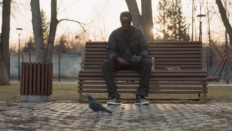man in dark jacket and cap sits on park bench at sunset throwing food to pigeon on paved ground, juice bottle and book beside him, with blurred person pushing stroller in background