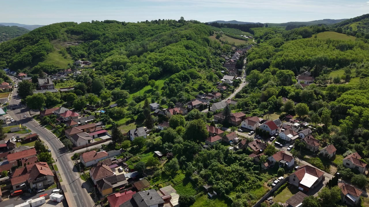 Widok z drona przedstawia spokojną węgierską wioskę położoną wśród bujnych, zielonych wzgórz pod jasnym błękitnym niebem.