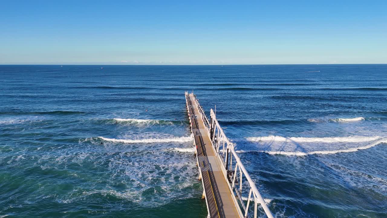 Drone footage captures a jetty extending into the ocean at Gold Coast, Australia, with waves gently rolling under clear blue skies