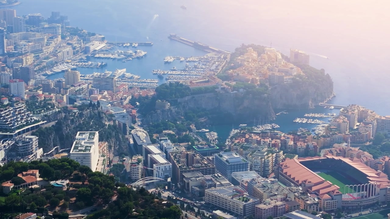Questa veduta mette in risalto il porto e la città di Monaco con i suoi edifici e la sua conformazione costiera. Si notano barche in acqua e strutture che costeggiano la riva in pieno giorno.