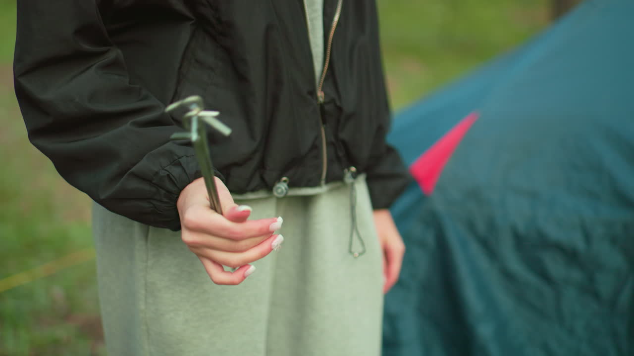 Close up of lady in black jacket and gray pants holding tent peg casually near tent during camping setup in forest, playfully turning metal peg in hand while standing on grassy outdoor terrain