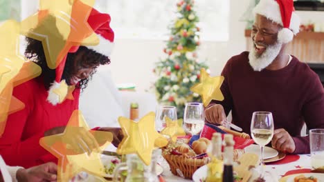 Yellow-star-falling-against-african-american-family-laughing-while-having-dinner-during-christmas