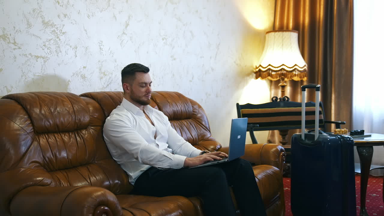 Handsome man with a laptop in cosy hotel room. Businessman sitting on a couch and working online at business trip. Travel suitcase in the room.