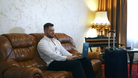handsome man with a laptop in cosy hotel room. businessman sitting on a couch and working online at business trip. travel suitcase in the room.