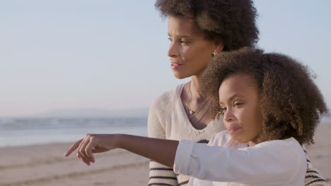 Cute-Girl-Talking-And-Pointing-Something-Into-Distance-While-Eating-Croissant-With-Her-Mom-During-A-Picnic-At-The-Beach