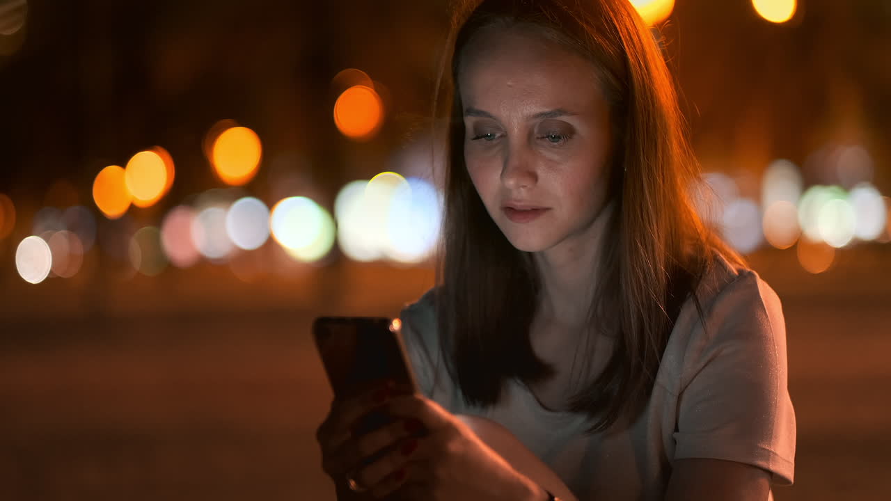 A beautiful young girl in the summer in a big night city keeps a smart phone in shell and writes a message reads e-mail and communicates in chat rooms and social networks keeps a blog looks at the phone screen