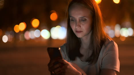 una hermosa joven en el verano en una gran ciudad nocturna mantiene un teléfono inteligente en la cáscara y escribe un mensaje lee correo electrónico y se comunica en salas de chat y redes sociales mantiene un blog mira la pantalla del teléfono