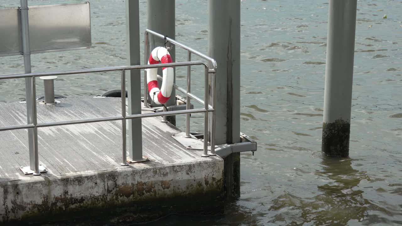 Detail of a canal dock or pier in Bangkok with a red-and-white lifebuoy attached to metal safety rails, pilings rising from the water and soft sunlight on the floating platform