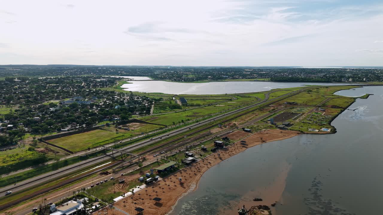 Aerial wide shot as a drone pulls away from the sandy Balneario Costa Sur and Paraná River waterfront in Posadas, Misiones, Argentina, with visible beach umbrellas, beachgoers, local bars