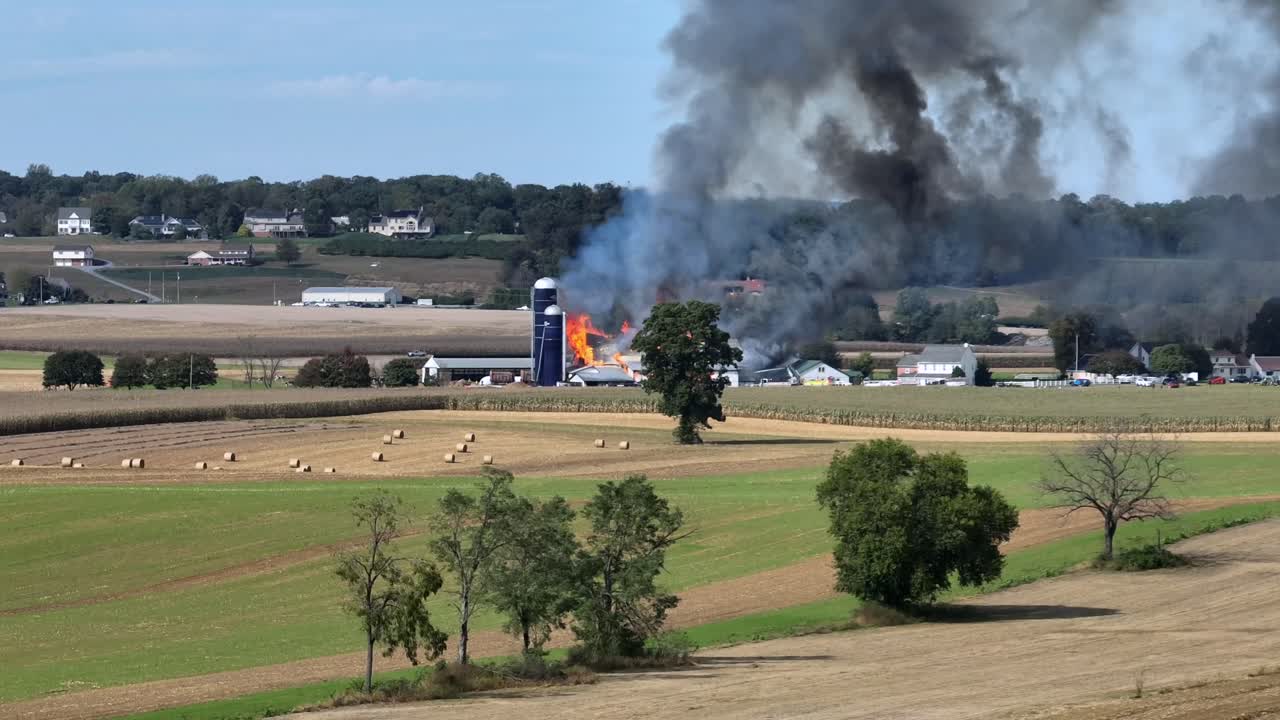Burning barn on rural American farmstead, smoke and ash rising over sunny fields. Fire truck and firefighters using water hoses to extinguish blaze. Dramatic countryside scene. Aerial wide shot