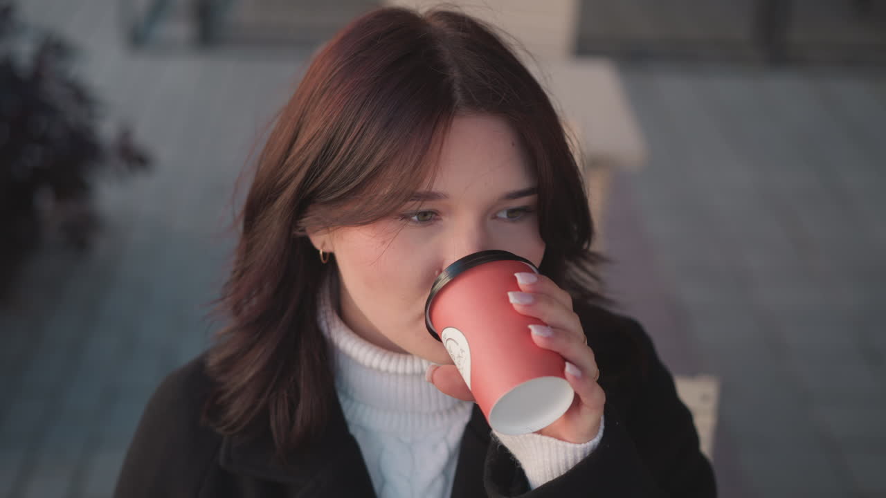 фрилансер в черном пальто и белой черепаховой шее глотает кофе вдумчиво, прежде чем осторожно опустить чашку, на заднем плане - деревянный стул и цветок