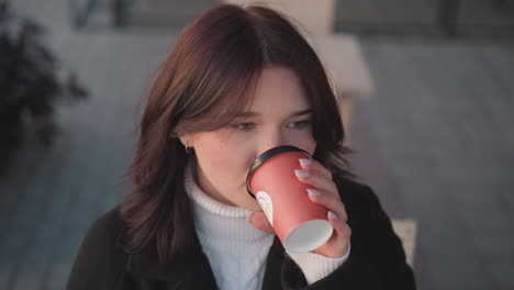 검은 코트와  거북이 목을 입은 프리랜서가 컵을 부드럽게 내려놓기 전에 커피를 조심스럽게 마시고, 배경에는 나무 의자와 꽃이 있습니다.