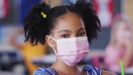 Portrait-of-african-american-schoolgirl-wearing-face-mask,-sitting-in-classroom-looking-at-camera