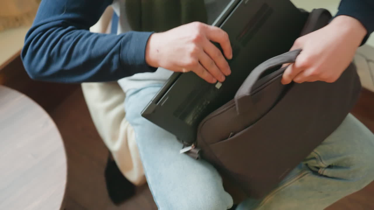 Portátil recogido en cafetería moderna, profesional concentrado cierra portátil delgado, guarda el dispositivo en mochila acolchada, asegura cremallera y correas, revisa asa y compartimentos, trayecto matutino tranquilo pero con propósito