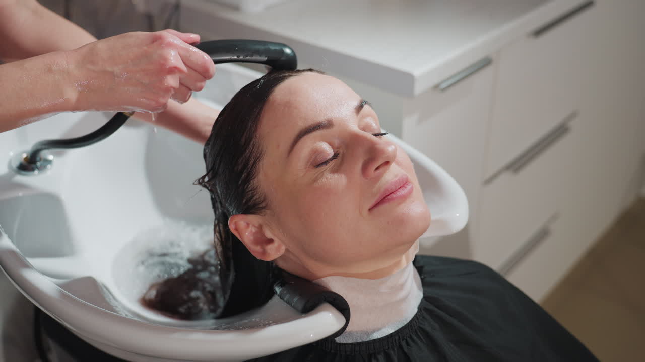 흰색으로 된 여자 리클라이닝, 스타일리스트가 휴대용 분무기를 사용하여 젖은 검은 머리를 감고, 따뜻한 물, 목 패드, 수건 깃, 눈을 감고, 차분한 표정, 현대적인 인테리어로 된 미용실 세면대에서 머리 헹구기