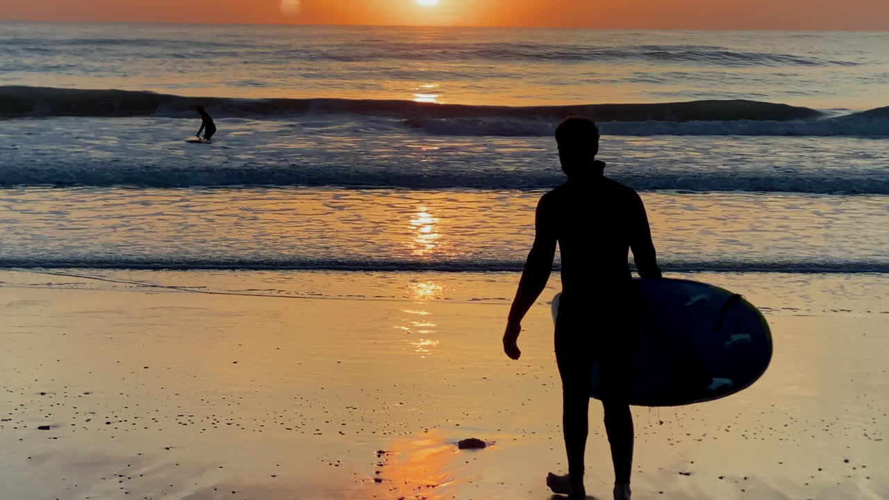 A lone surfer walking toward the glowing horizon across reflective wet sand a dramatic orange sunset sky over the wide sandy beach of Costa Ballena with calm gentle waves