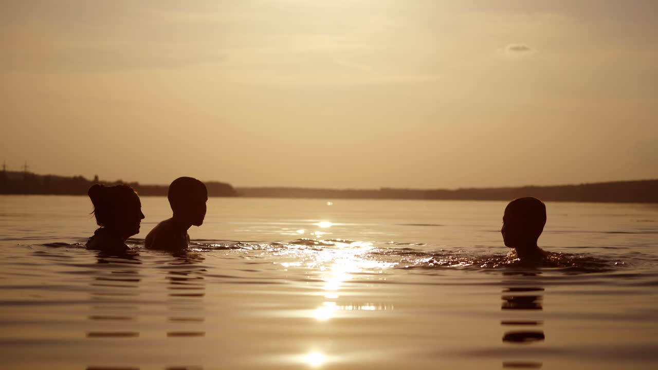 Woman and two children playing in water in the evening. Silhouette of female and boys having fun in the river at sunset. Mother throws up her son into the water.