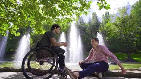 Young-disabled-man-sitting-in-a-wheelchair-talking-to-his-friend-outdoors.