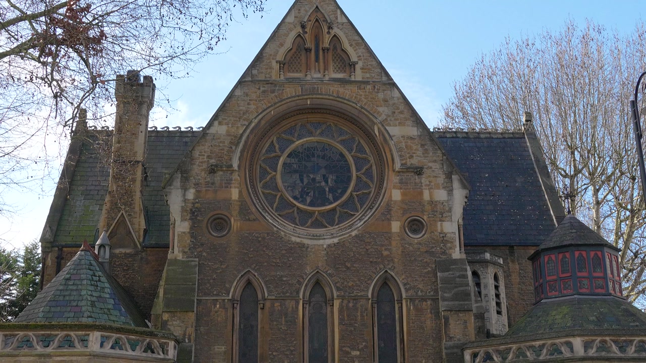 Front view of St Mary Abbots Church in London featuring its distinctive Gothic architecture and large circular rose window. The historic facade is framed by trees, showcasing detailed stonework