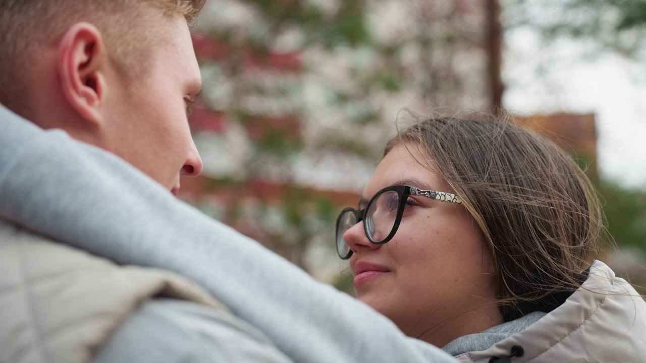 Close up of young lovers gazing at each other as man tenderly adjusts woman hair while wind gently flutters her strands, sharing affectionate moment with deep eye contact and warmth outdoors
