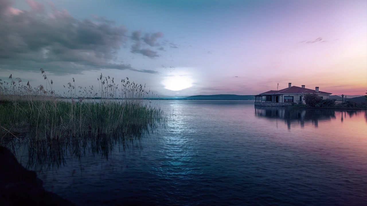 Fredelig solnedgang over Balatonsøen med roligt reflekterende vand, siv ved søbredden og et hus ved vandet under en farverig tusmørkehimmel i Ungarn.