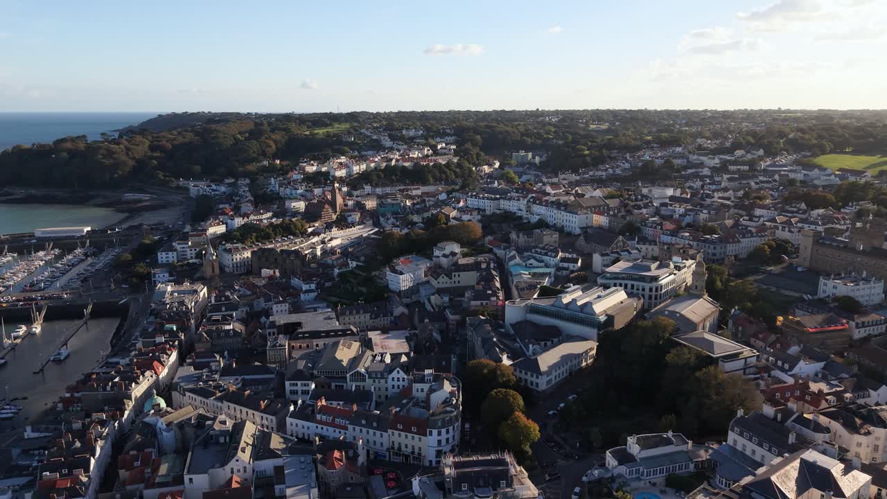 Aerial footage of St Peter Port Guernsey including harbour and Hevelet Bay high but detailed flying south in later afternoon sun with wooded cliffs and sea in the distance