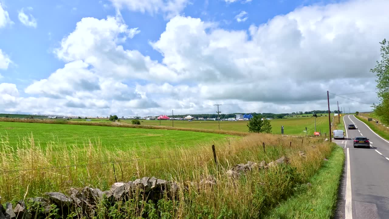 A vehicle travels along a scenic rural road bordered by fields and stone walls under bright daylight, passing festival signage and temporary surveillance equipment