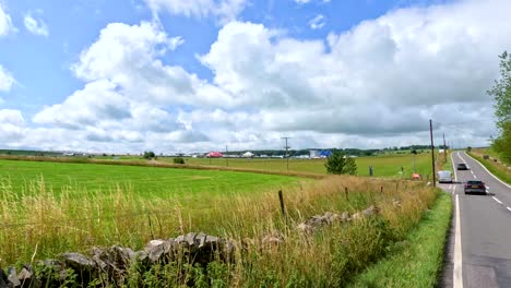 a vehicle travels along a scenic rural road bordered by fields and stone walls under bright daylight, passing festival signage and temporary surveillance equipment