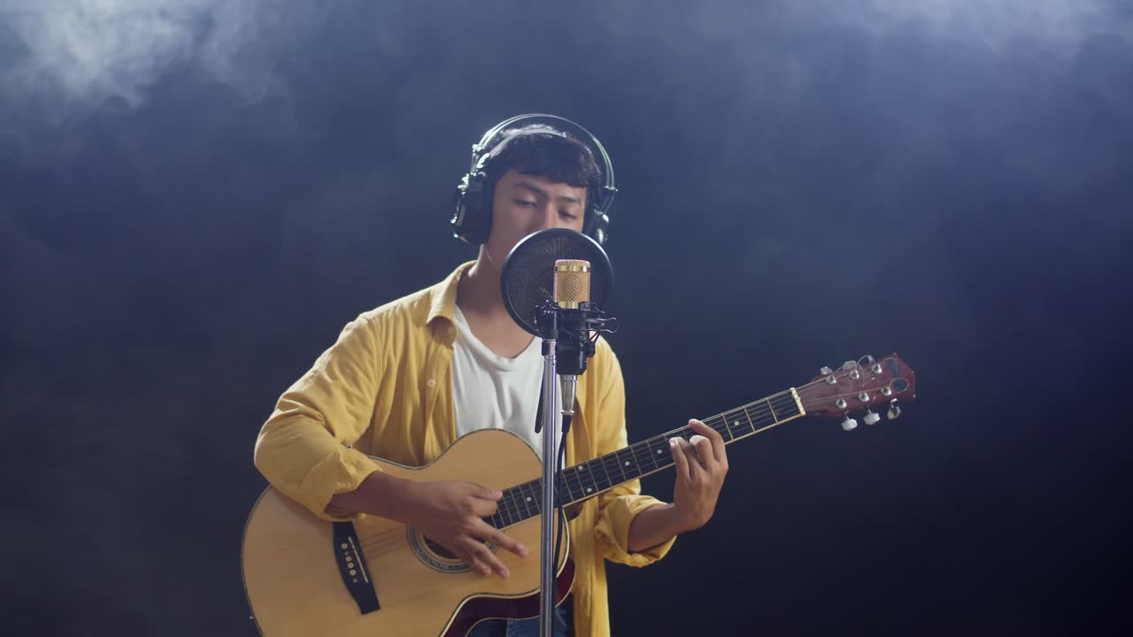 헤드폰을 들고 있는 아시아 소년이 기타를 연주하고 검은색 배경의  연기에 콘덴서 마이크로 노래하고 있다