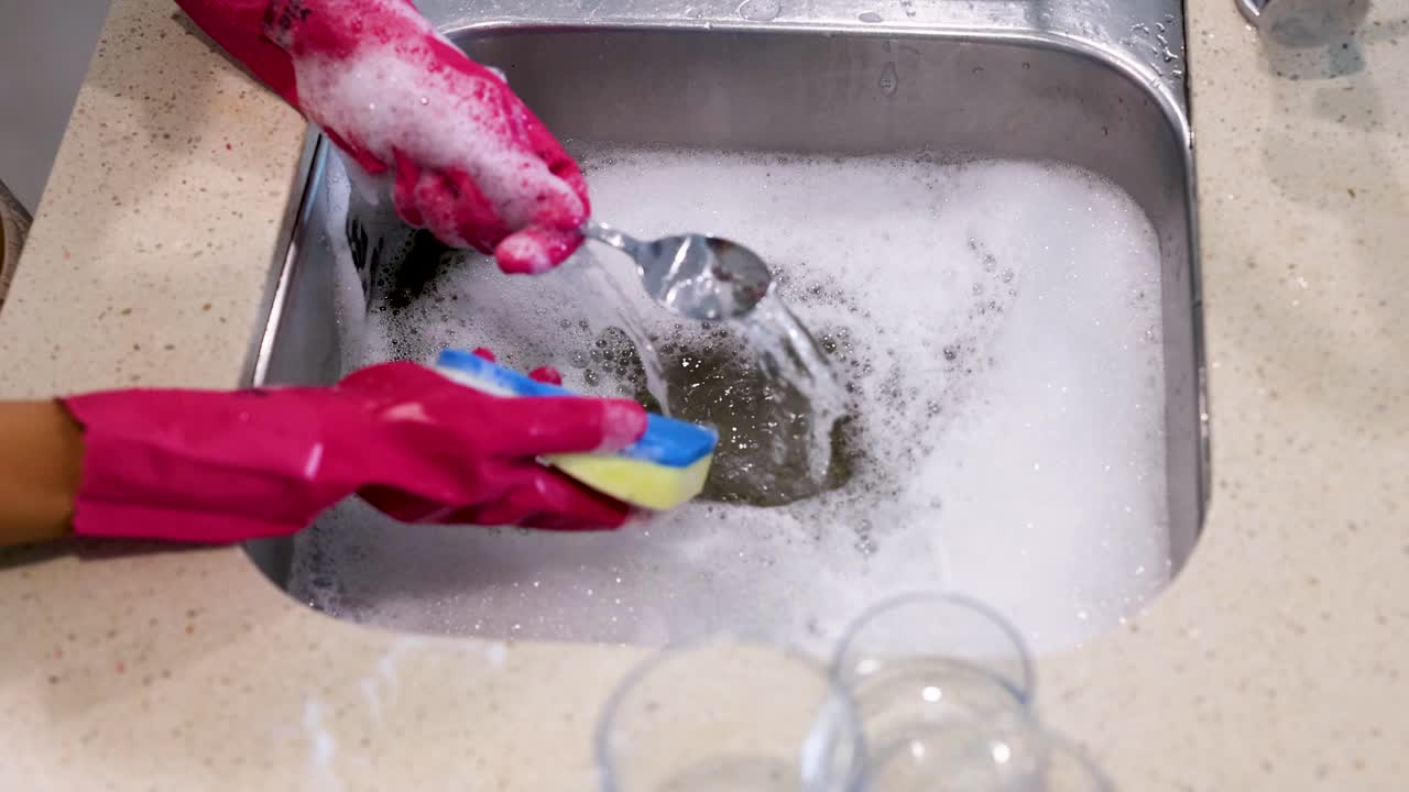 Hands wearing pink gloves wash a spoon in a soapy kitchen sink. Bright lighting highlights the cleaning process