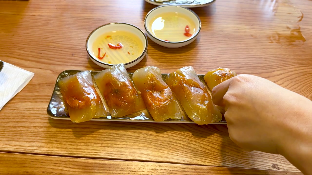 A person picks up a translucent shrimp dumpling from a platter on a wooden table
