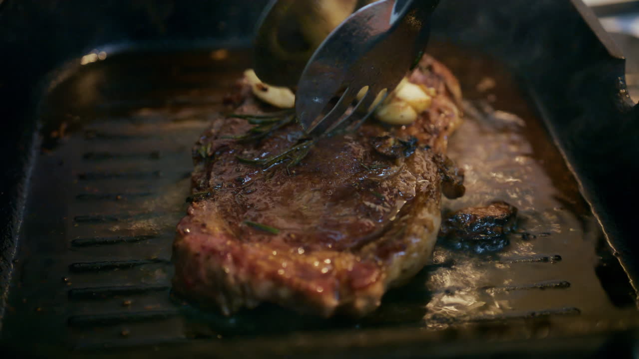 Nahaufnahme eines saftigen Steaks, das in einer Grillpfanne brutzelt, während es mit einem Wender angedrückt wird.