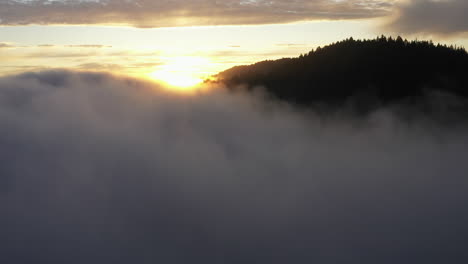 drone flight through fog revealing evergreen covered mountains and a golden sunset