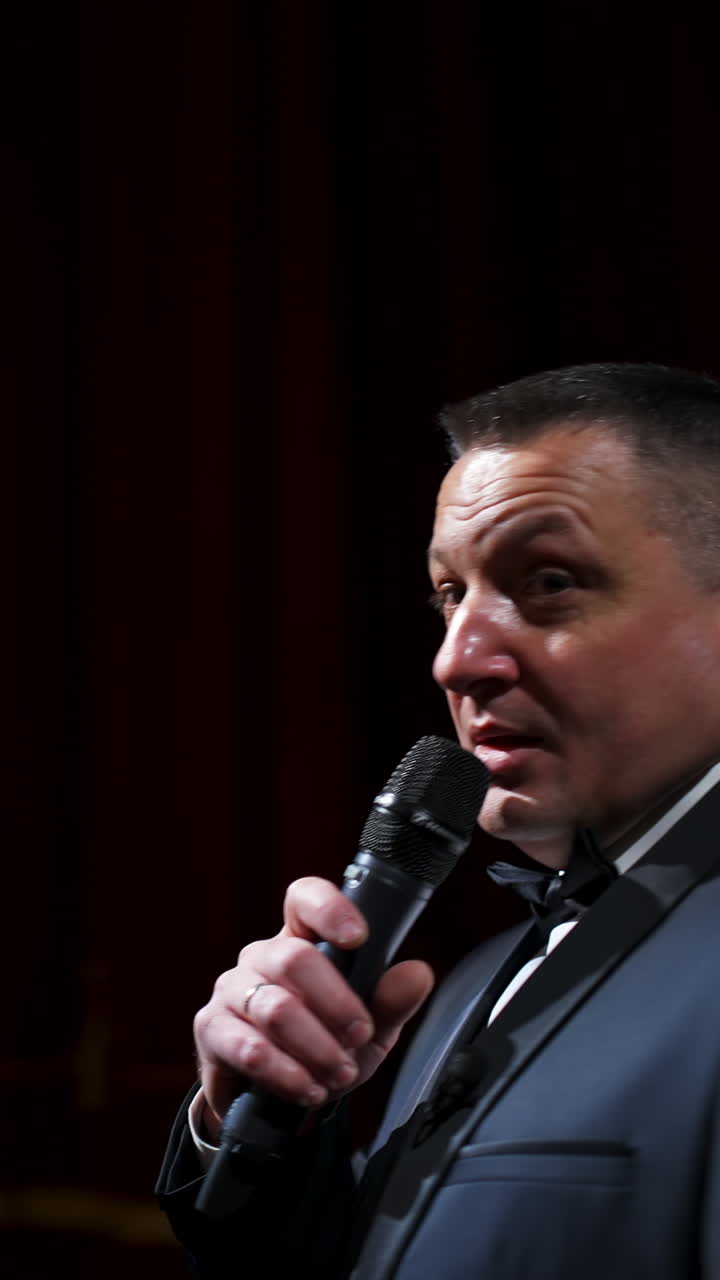 Experienced host with a microphone. Positive man in gray costume standing on theatrical stage and talking on camera. Lockdown during pandemic. Vertical video