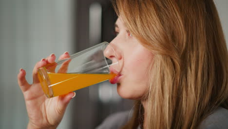close up side view of woman with long hair sipping orange juice from glass cup in kitchen, natural light highlights her smooth skin and healthy lifestyle
