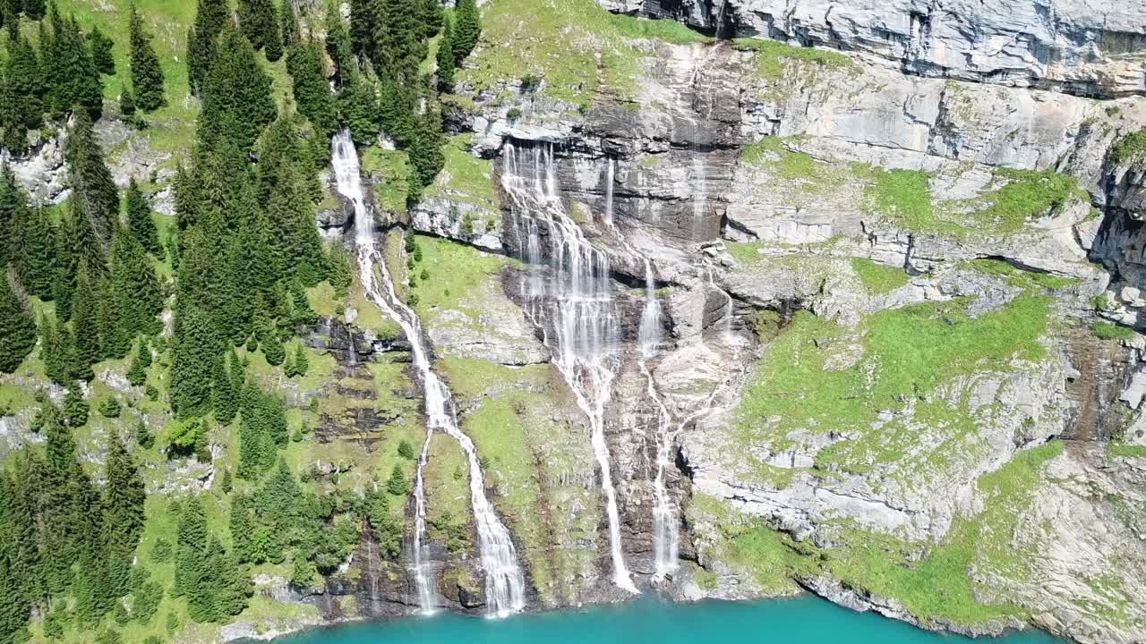 Multiple waterfalls are descending from a steep cliff covered with lush green vegetation, flowing into a turquoise lake, creating a breathtaking landscape in the swiss alps, Oeschinensee, Switzerland