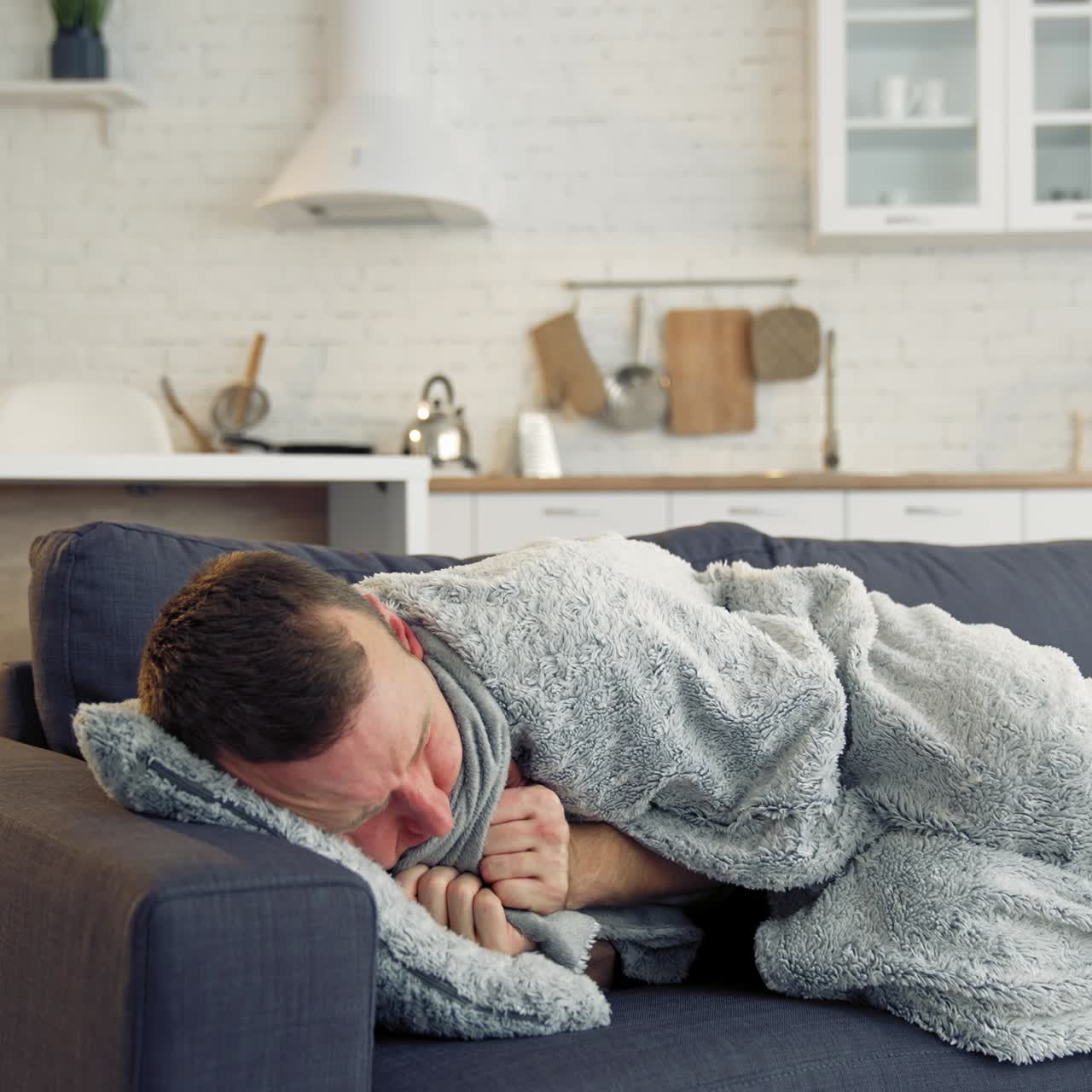 Young man trembling from fever. Sick man lying on couch at home and covering with warm blanket over the head