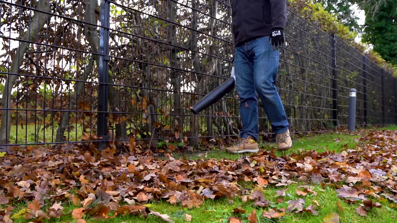 Man blowing leaves with leaf blower next to fence in slow motion