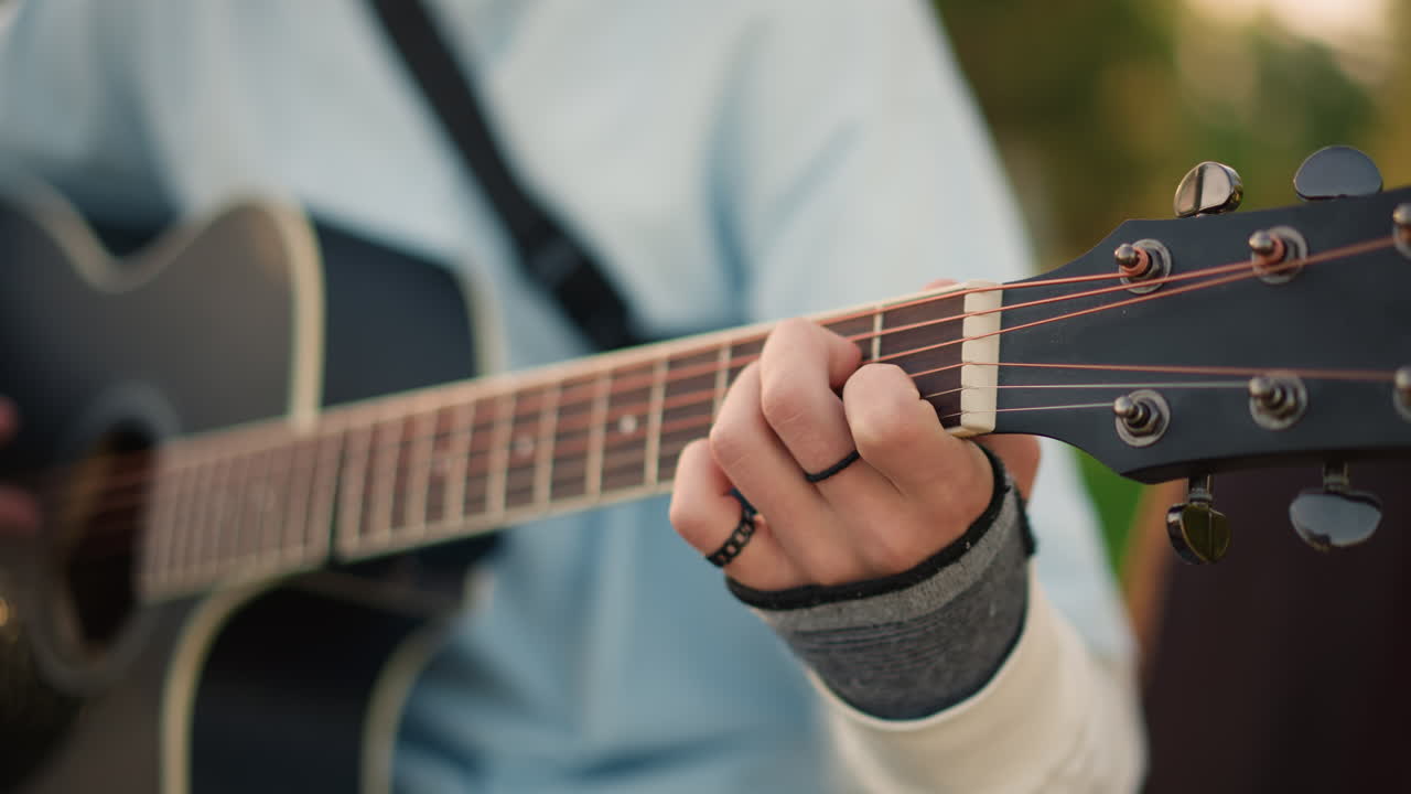 closeup left hand fretting guitar headstock outdoors, sleeve and ring detail, tuning pegs and worn strings, warm bokeh and soft autumn light, songwriter practicing chords on park bench, calm focused