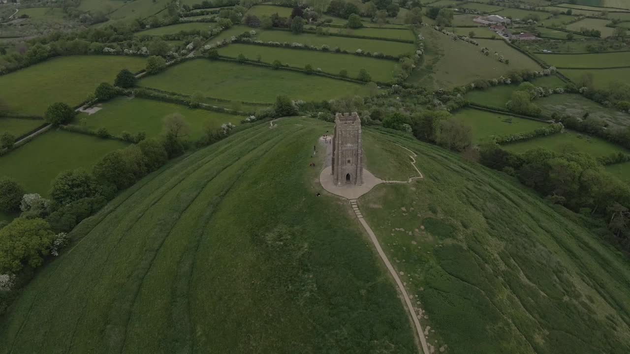Aerial view of Glastonbury, drone rotating and camera facing down over the Glastonbury Tor, drone flying over the green fields with some houses on the left side of the camera. 4K, 60fps.