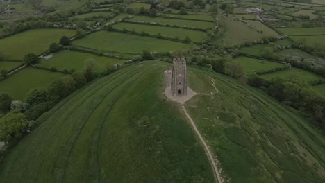 aerial view of glastonbury, drone rotating and camera facing down over the glastonbury tor, drone flying over the green fields with some houses on the left side of the camera. 4k, 60fps.