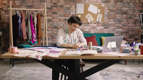 en el taller de moda y costura delante de la cámara increíble aspecto mujer diseñadora de moda coser algunas ropas luego mirando hacia la cámara y sonriendo ampliamente
