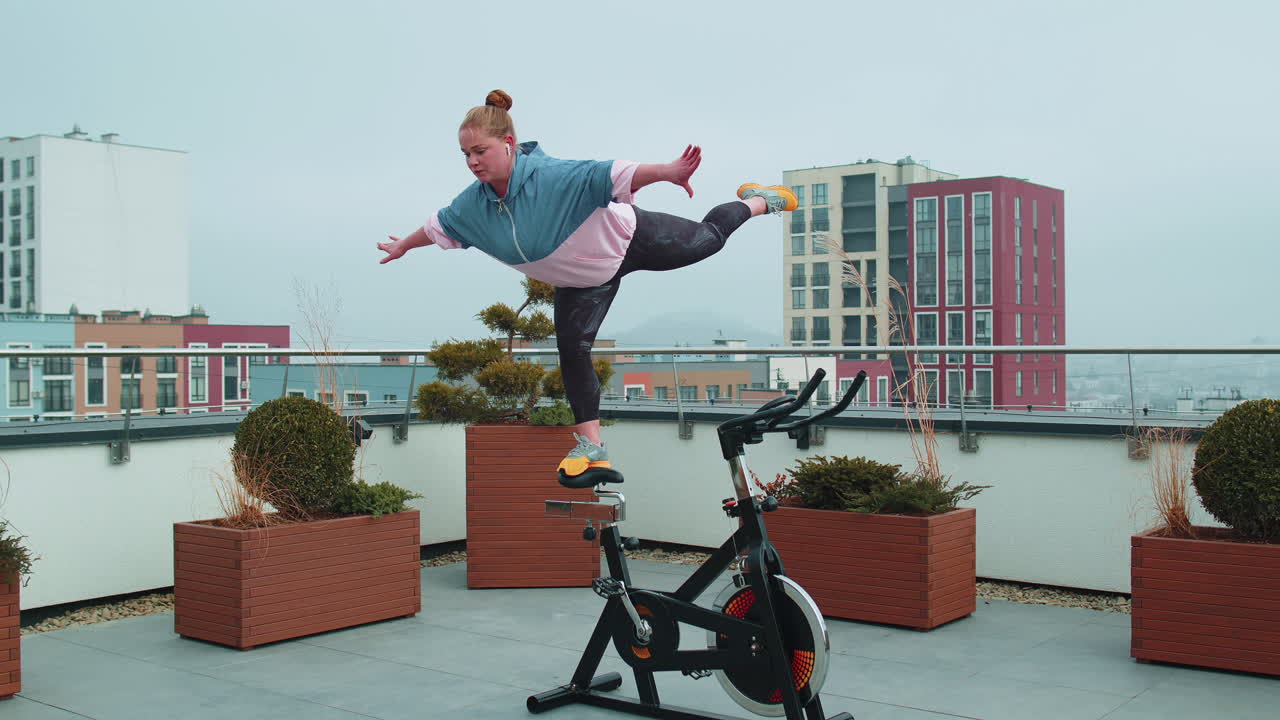 Athletic girl performing swallow acrobatic trick exercises on cycling stationary bike on house roof