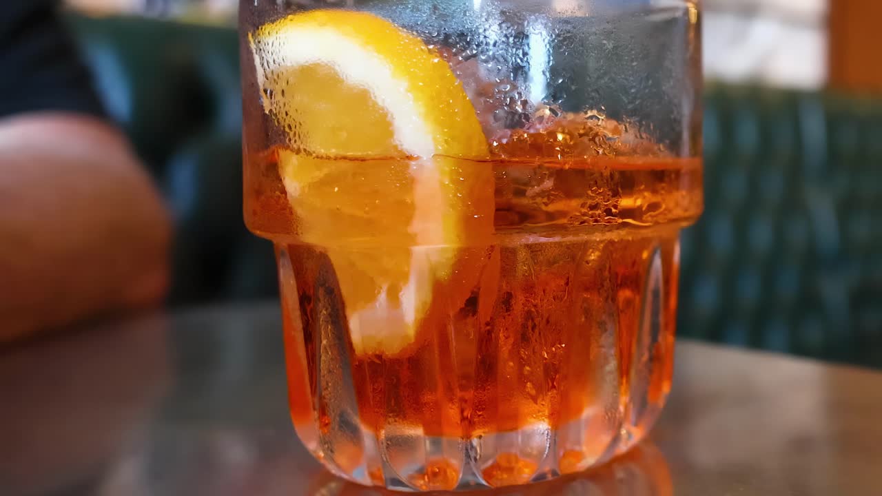 Un primo piano di un cocktail arancione con una fetta, posto su un tavolo riflettente.