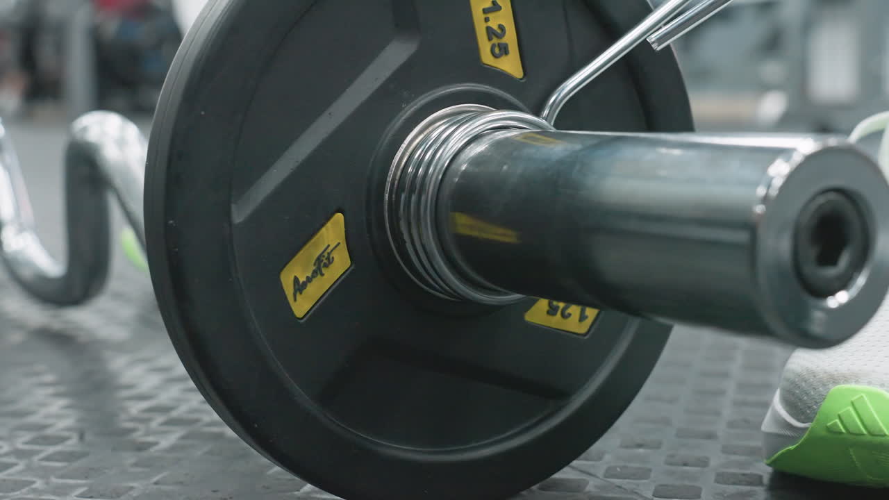 Close up of person clipping weight plate onto bar with foot and leg visible in gym group on rubber floor, showcasing hands securing metal spring collar on chrome bar end during workout prep