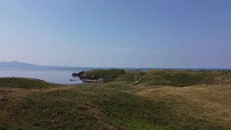 Vista-Aérea-De-La-Isla-De-Llanddwyn,-Sendero-Para-Caminar-Hacia-El-Faro-De-Piedra-Del-Faro-Y-Las-Brumosas-Montañas-De-Snowdonia