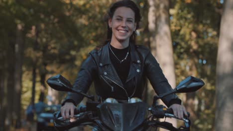 beautiful closeup of smiling white female on motor bike driving in leather jacket riding in forest, trees with golden autumn leaves color on sunny day